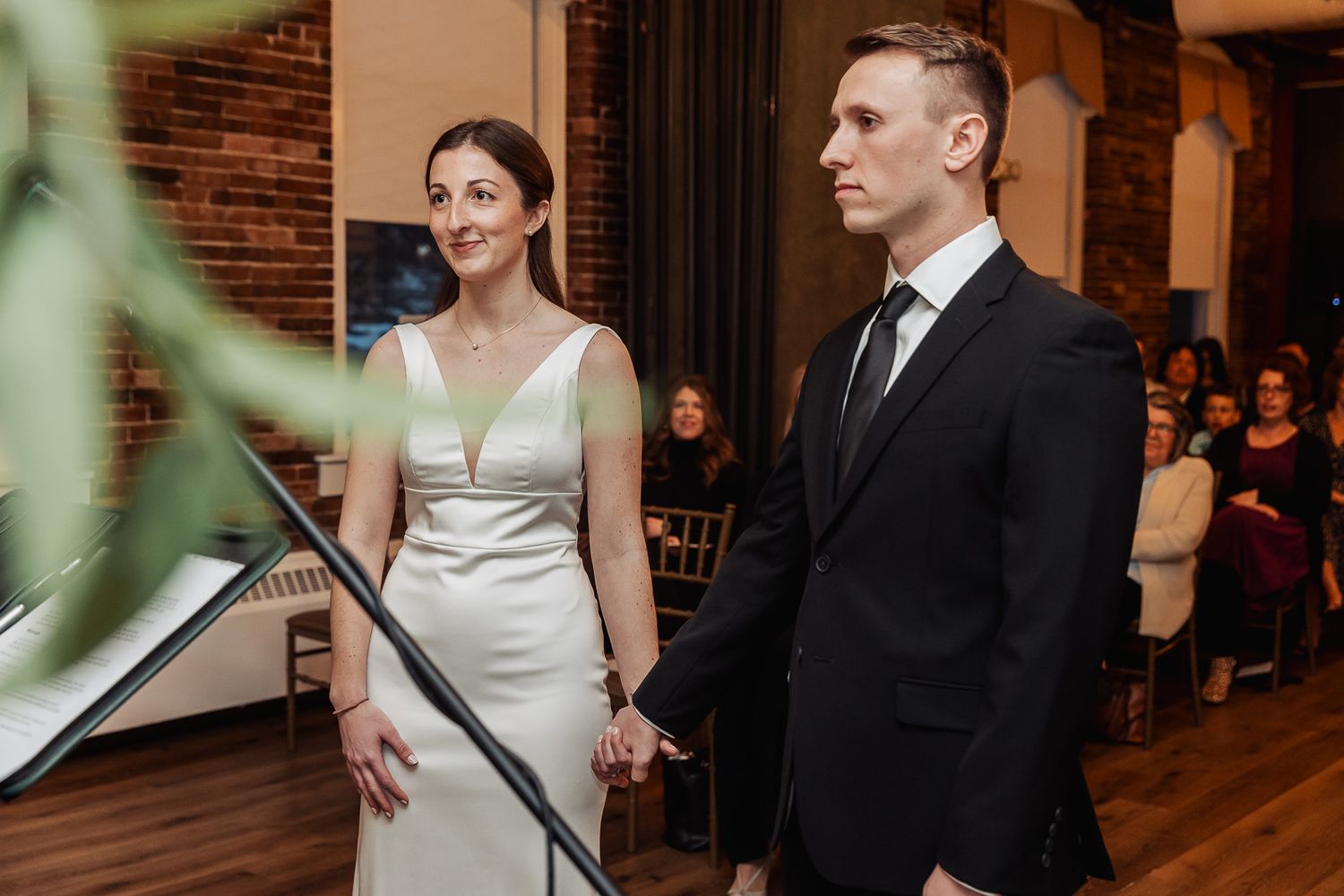 A couple stands together at a wedding ceremony in an elegant indoor venue with brick walls and wooden floors.