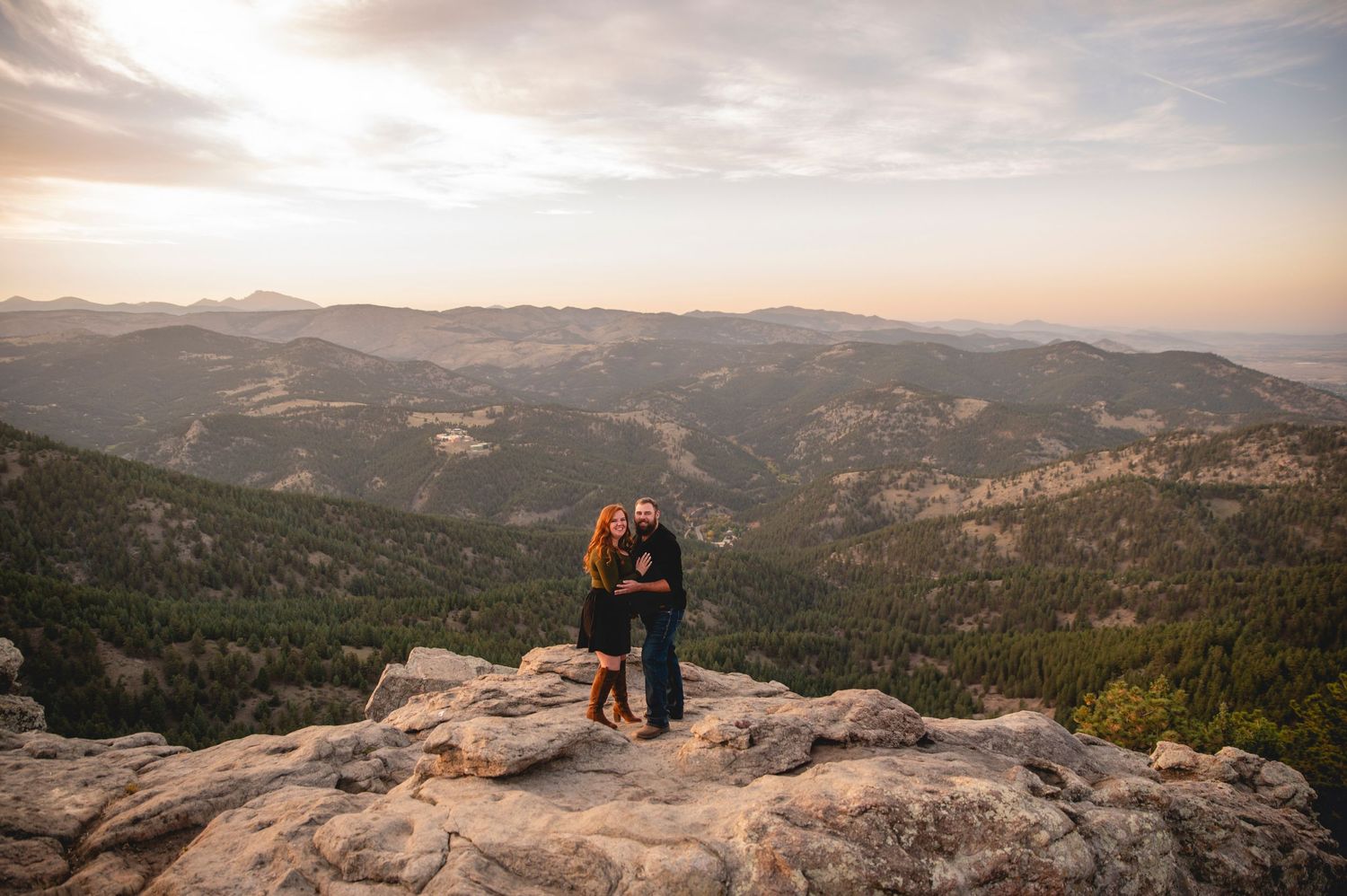 A couple embraces on mountain peak overlooking vast mountainous landscape at sunset.