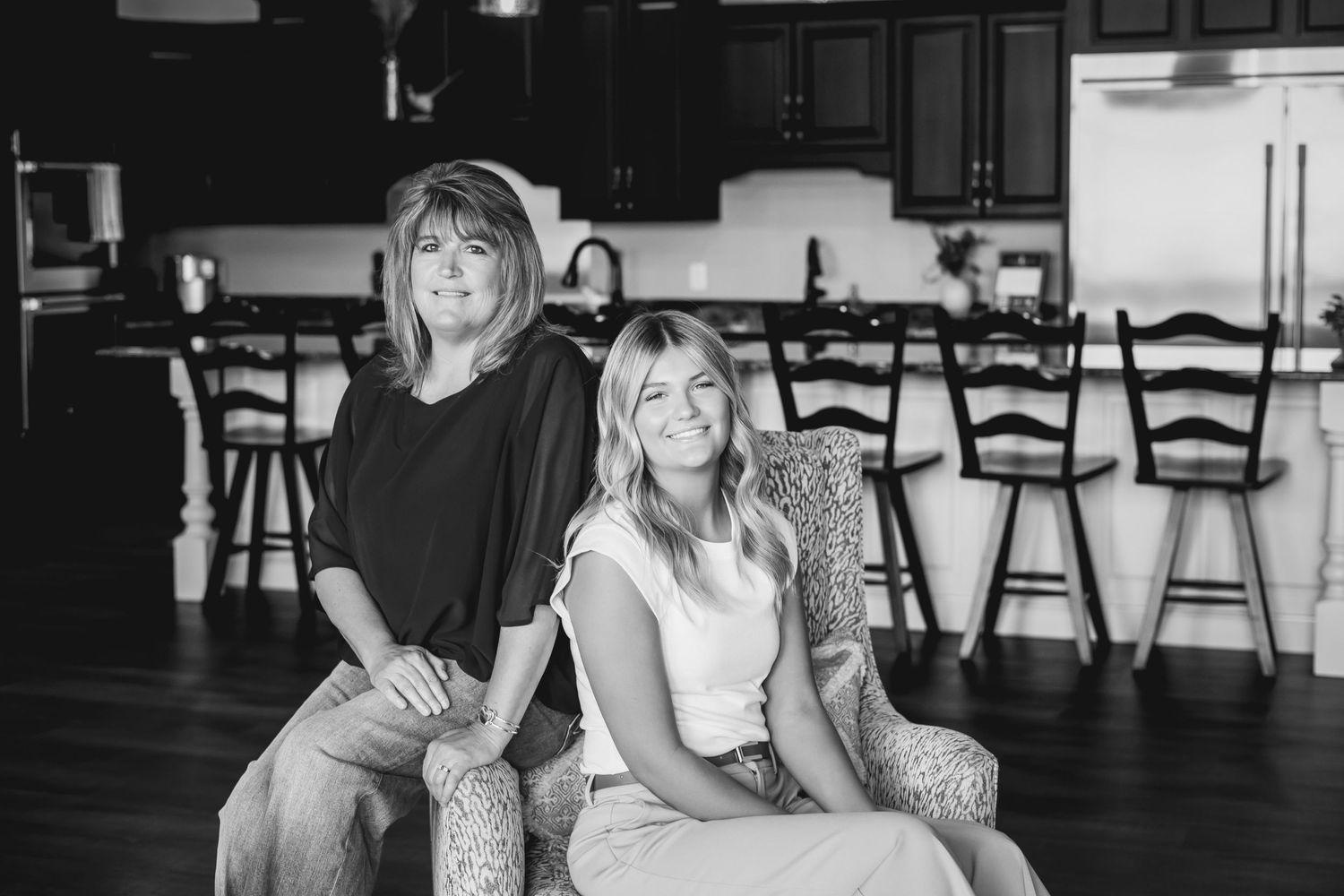 Two people pose together on furniture in a modern kitchen with dark hardwood floors and black bar chairs in background.