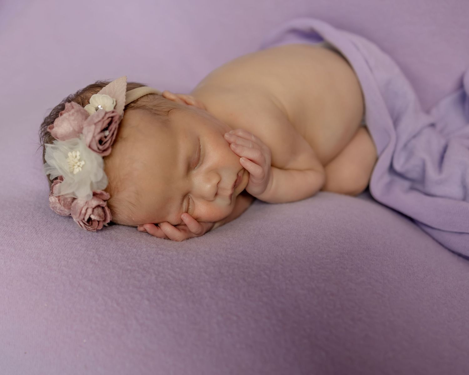 Sleeping newborn with floral headband on purple backdrop.