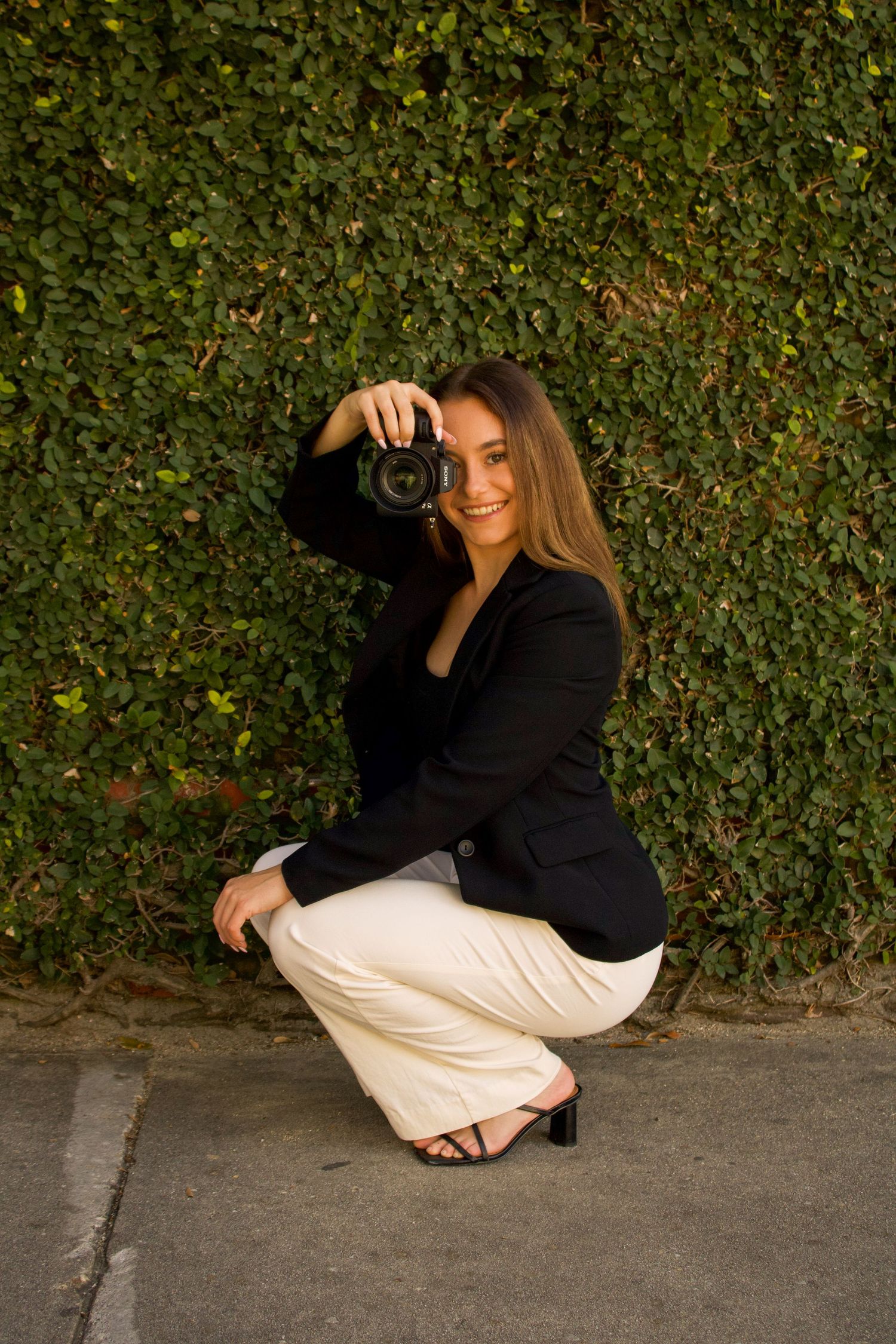 Photographer crouching in black blazer and white pants takes photos against green hedge wall in warm sunset light.
