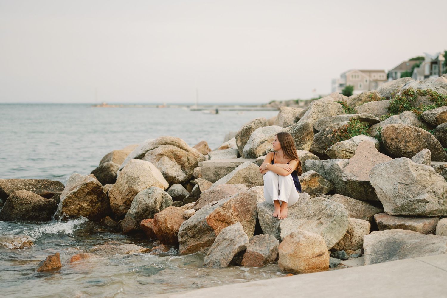 A woman in a white dress sits on rocky shoreline boulders with calm ocean waters and sailboats in the background.