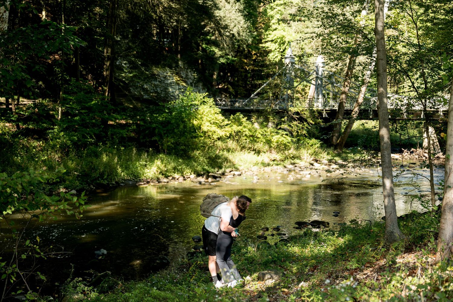Child exploring the edge of a serene forest stream surrounded by lush green trees and a wooden bridge in the background.