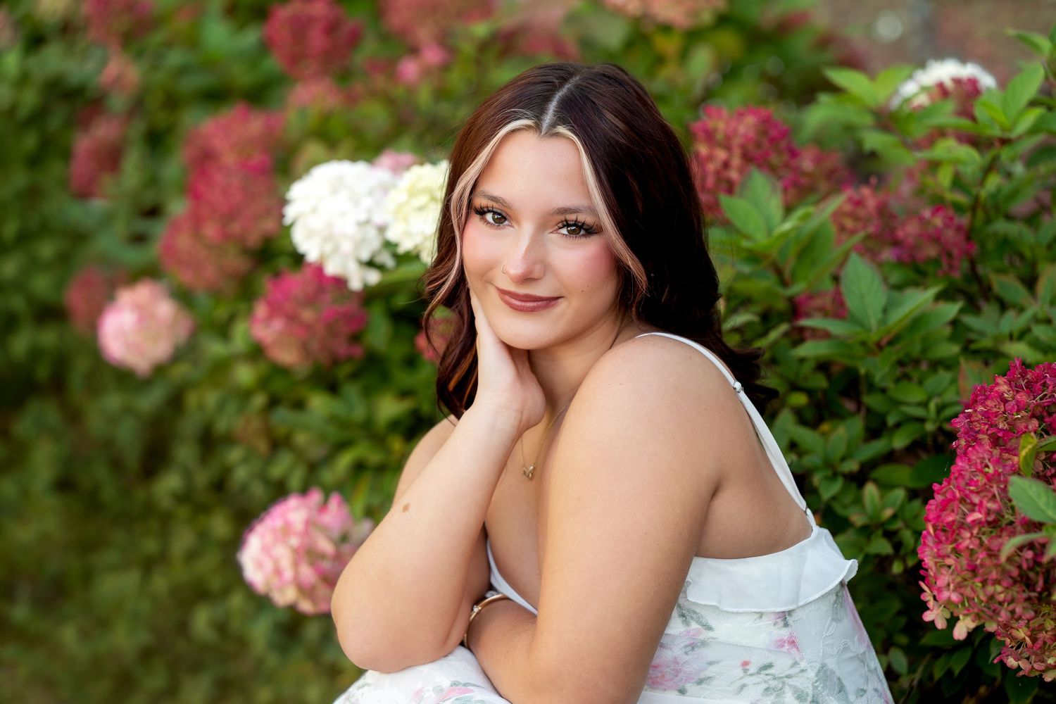 Young woman in floral dress poses gracefully among pink and white hydrangeas in a lush garden setting.