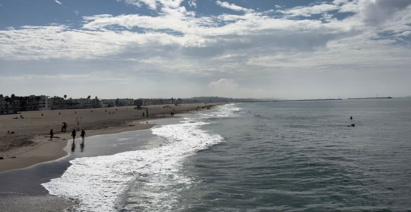 Panoramic view of a sandy beach with gentle ocean waves, scattered beachgoers, and a partly cloudy sky overhead.