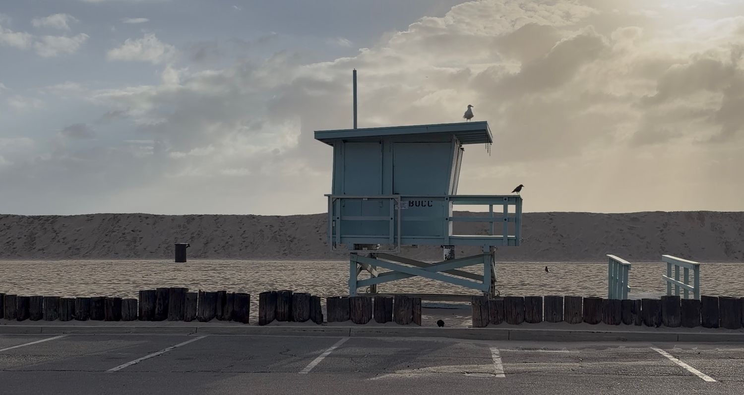 A blue lifeguard tower stands alone on an empty beach parking lot under a dramatic cloudy sky at dusk.