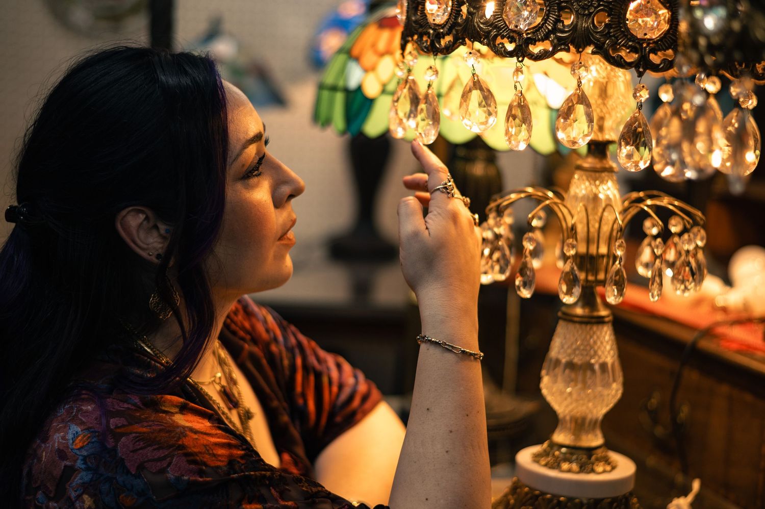 Woman examining an ornate crystal lamp with warm glowing light in a dimly lit antique shop.