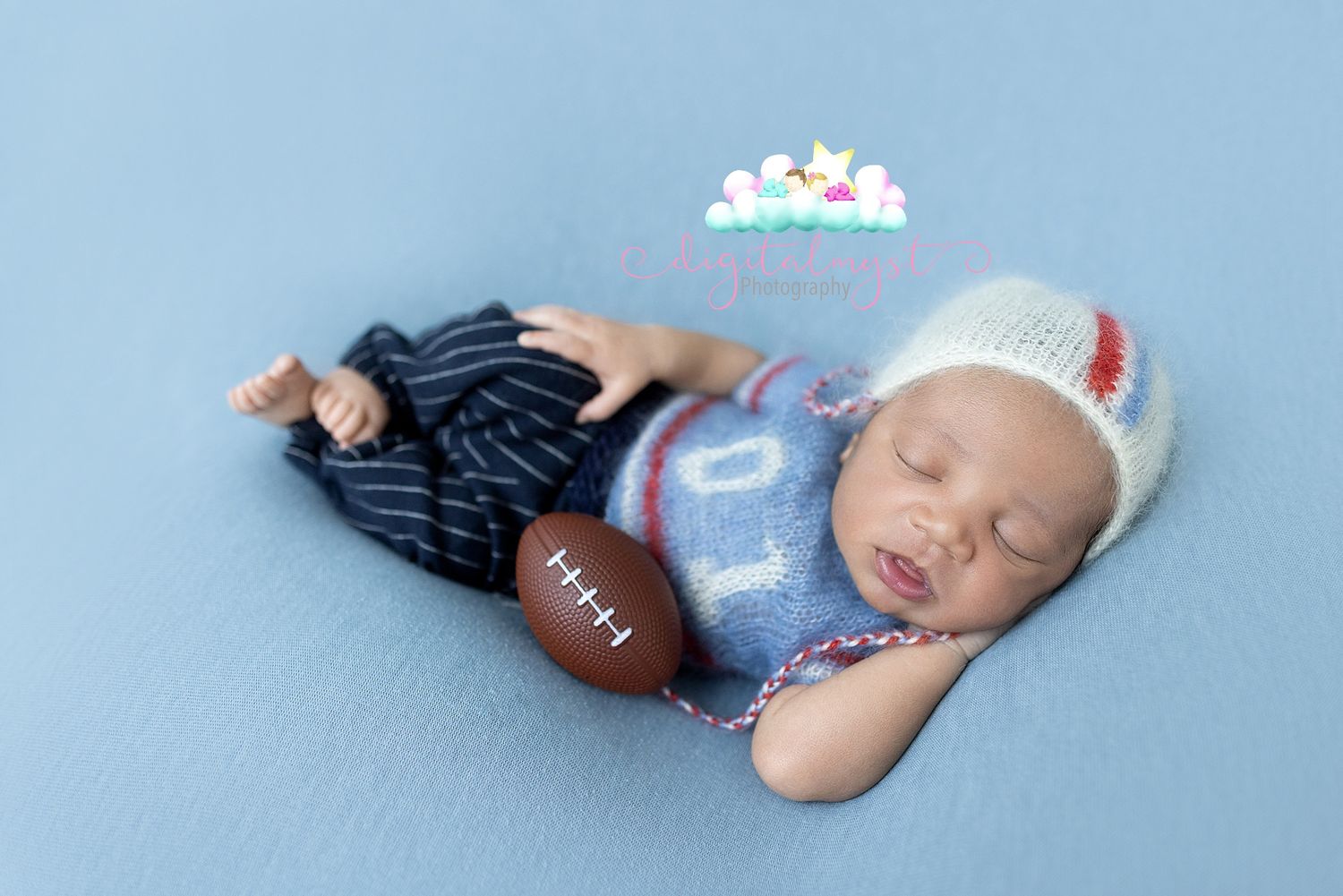 Newborn baby sleeping on blue blanket wearing knit hat holding a small football in blue striped outfit.