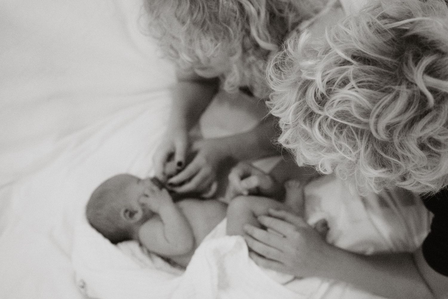 Black and white photo of a curly-haired woman tenderly gazing at a newborn baby lying on a white blanket.