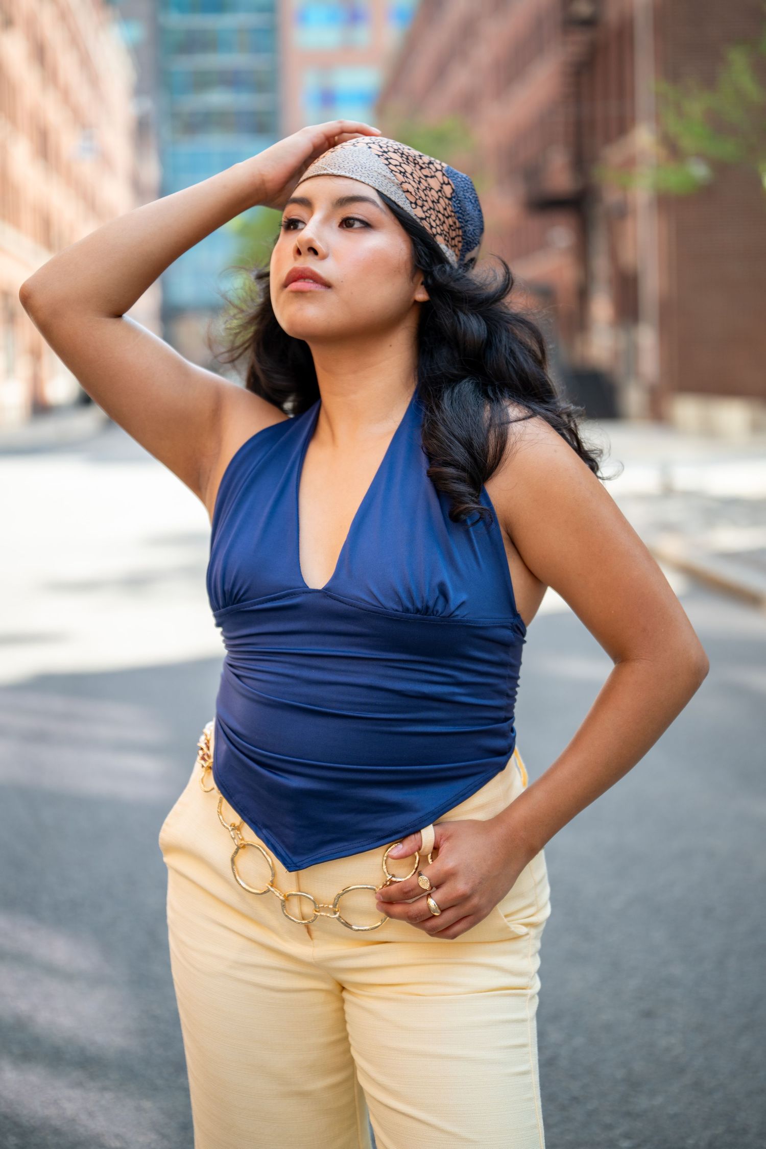 Woman in blue halter top, cream pants, and patterned headband poses on urban street.