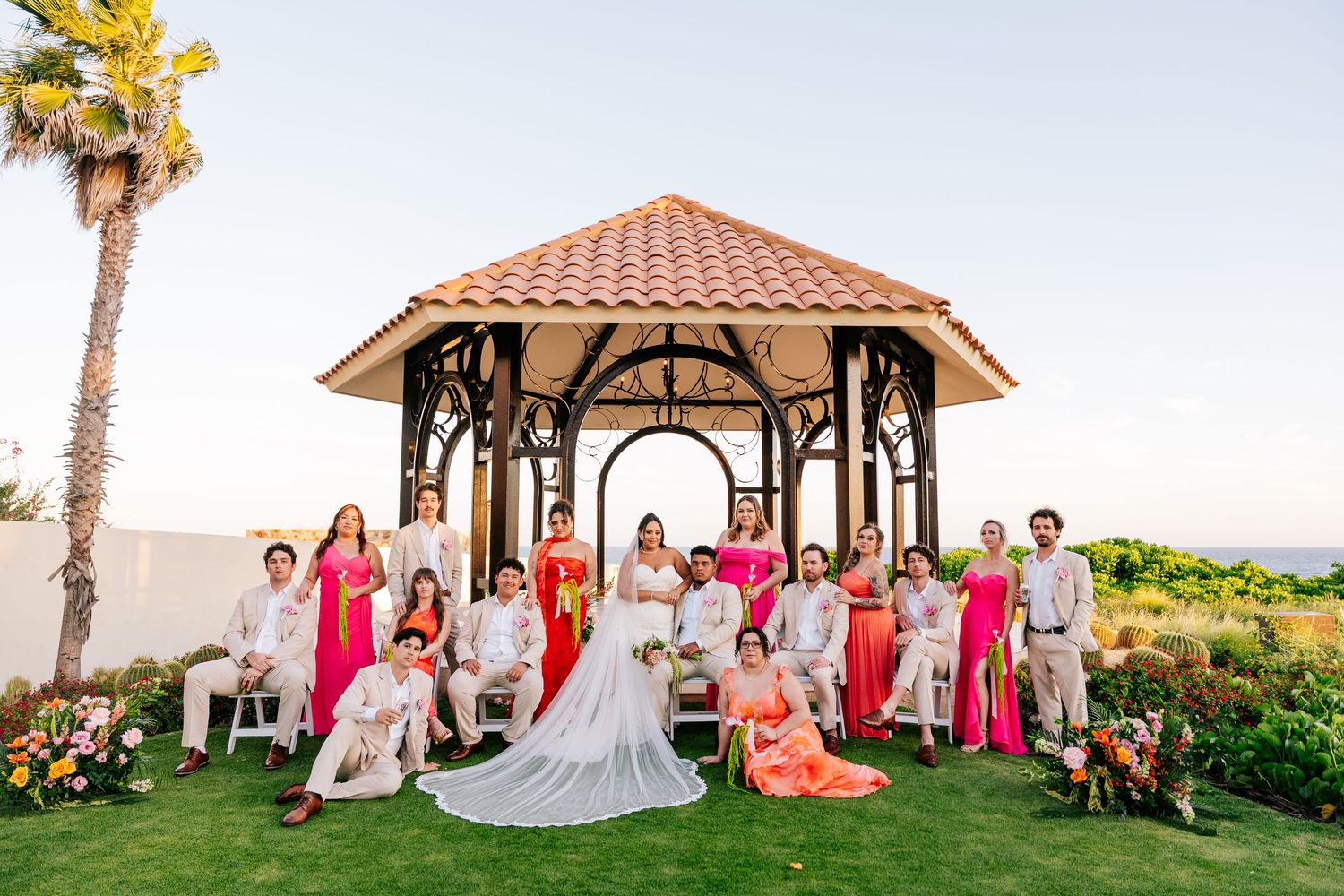 Bride in a long white gown poses with bridesmaids in pink and coral and groomsmen in light suits beneath a wooden gazebo by the ocean.