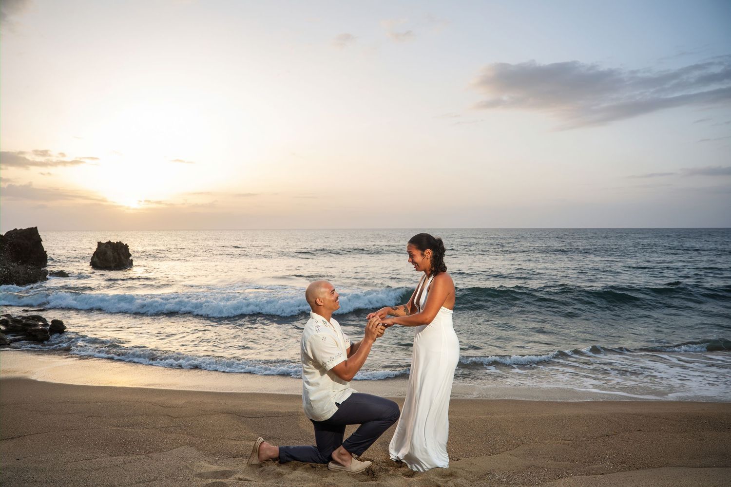 Man proposes to woman on sandy beach at sunset with ocean waves and golden sky in background.