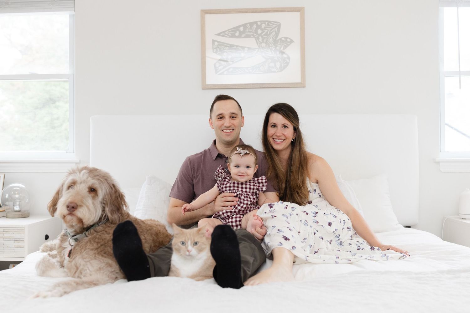 Family portrait of couple with baby, fluffy dog, and white cat sitting together on a white bed.
