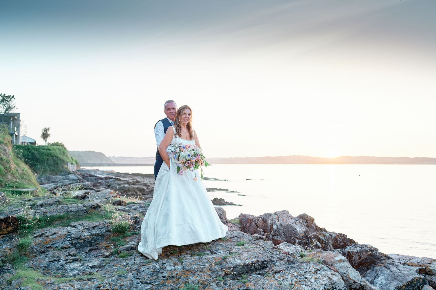 Bride and groom posing on rocky shoreline at sunset, bride in white gown holding bouquet, scenic ocean backdrop.