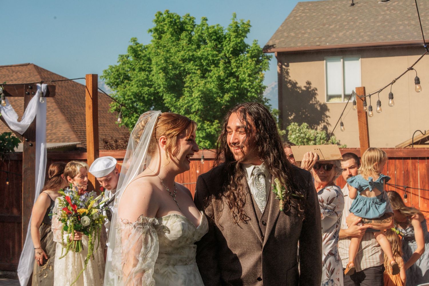 Newlywed couple smiling at each other during outdoor backyard wedding ceremony, bride holding colorful bouquet.