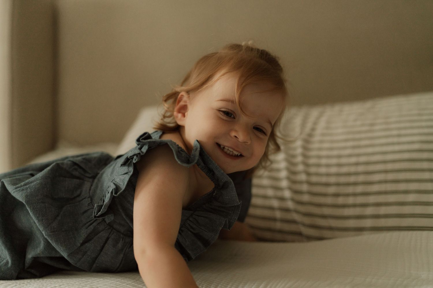 Smiling baby girl in a gray ruffle dress lying on a couch, looking happy and playful.
