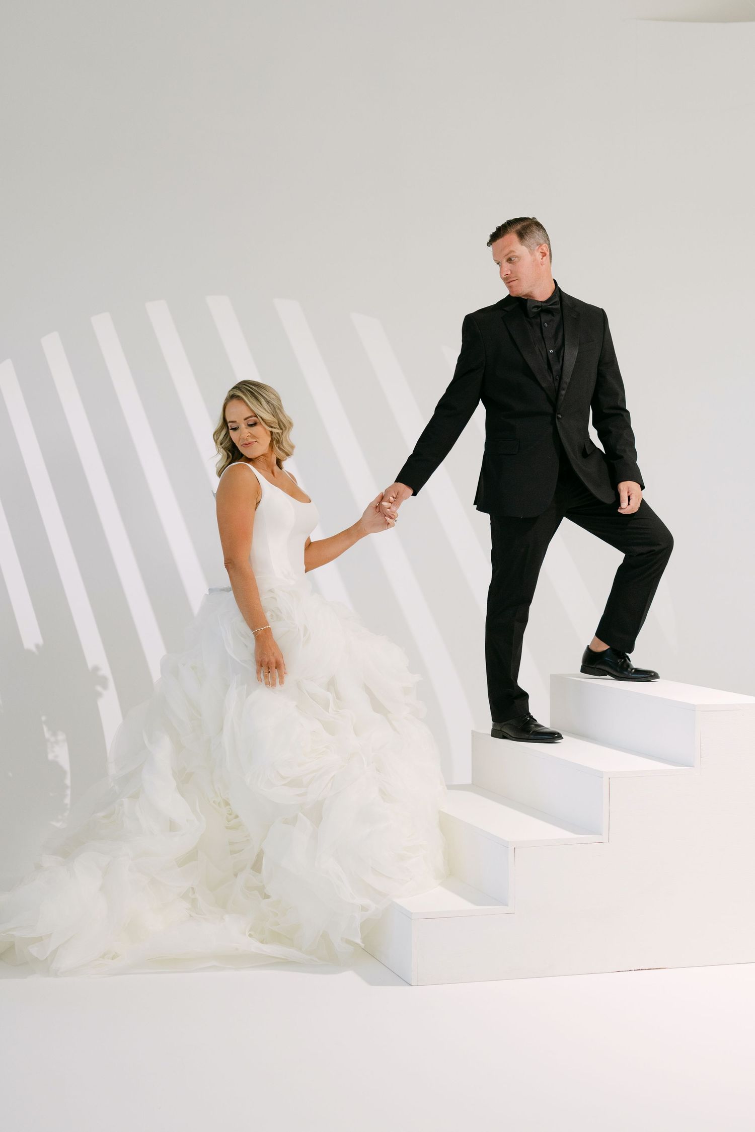 Bride in white ruffled gown and groom in black tuxedo hold hands descending white modern stairs.