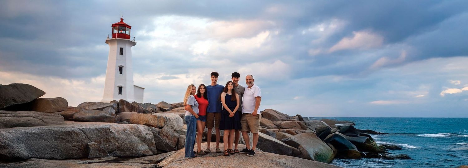 Family posing on rocky shore in front of Peggy's Cove lighthouse, Nova Scotia, under dramatic cloudy sky.