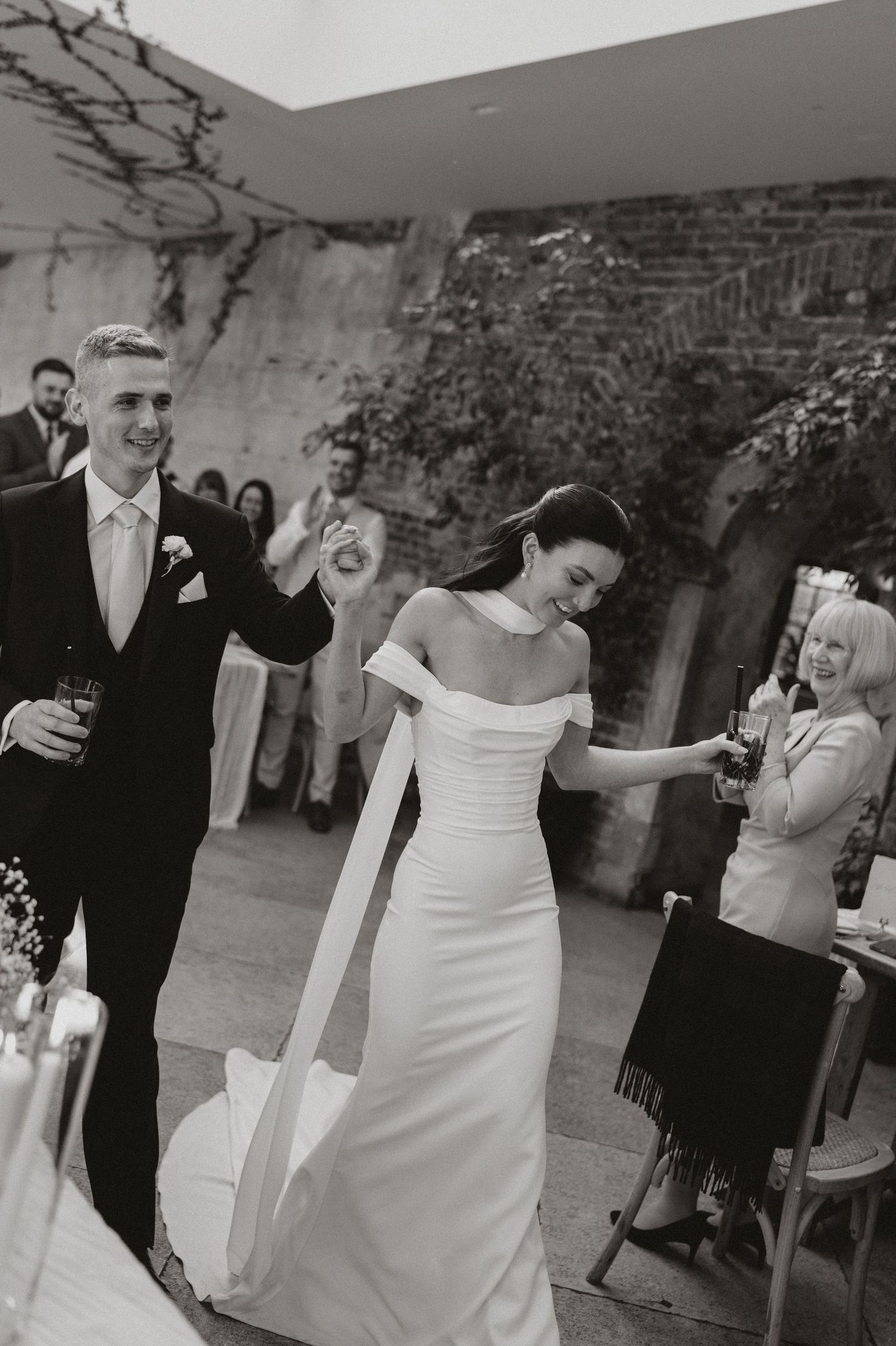 Black and white photo of a bride in off-shoulder gown and groom in suit making grand entrance at wedding reception.