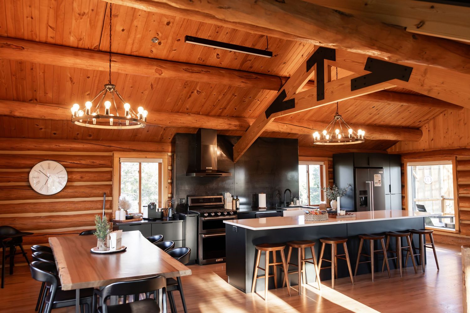 Rustic log cabin with wrap-around porch surrounded by pine trees under a clear blue sky on a gravel lot.