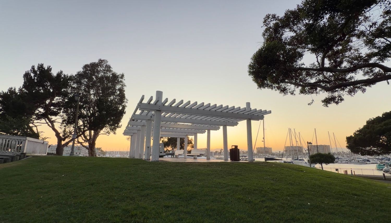 White pergola overlooking a marina at sunset, surrounded by lush green lawn and trees in a coastal park.