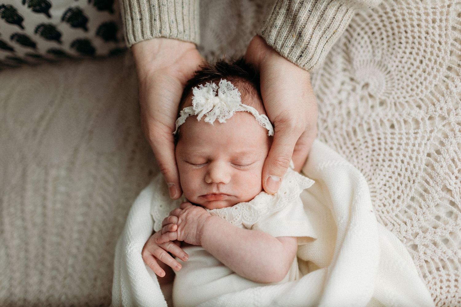 Sleeping newborn baby girl in white wrap with floral headband, cradled gently by parent hands.