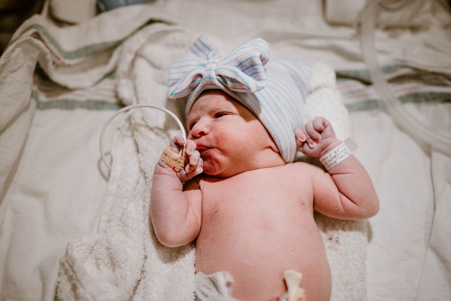 A newborn baby lies on white bedding while sucking their thumb.