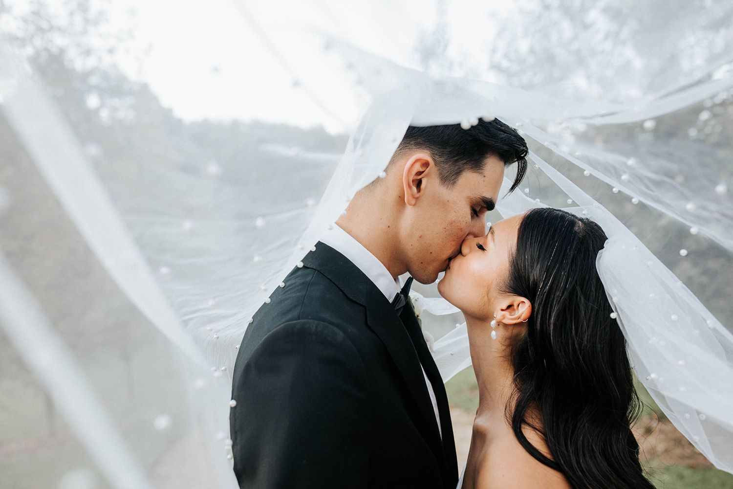 A bride and groom share a romantic kiss under a flowing white veil on their wedding day.