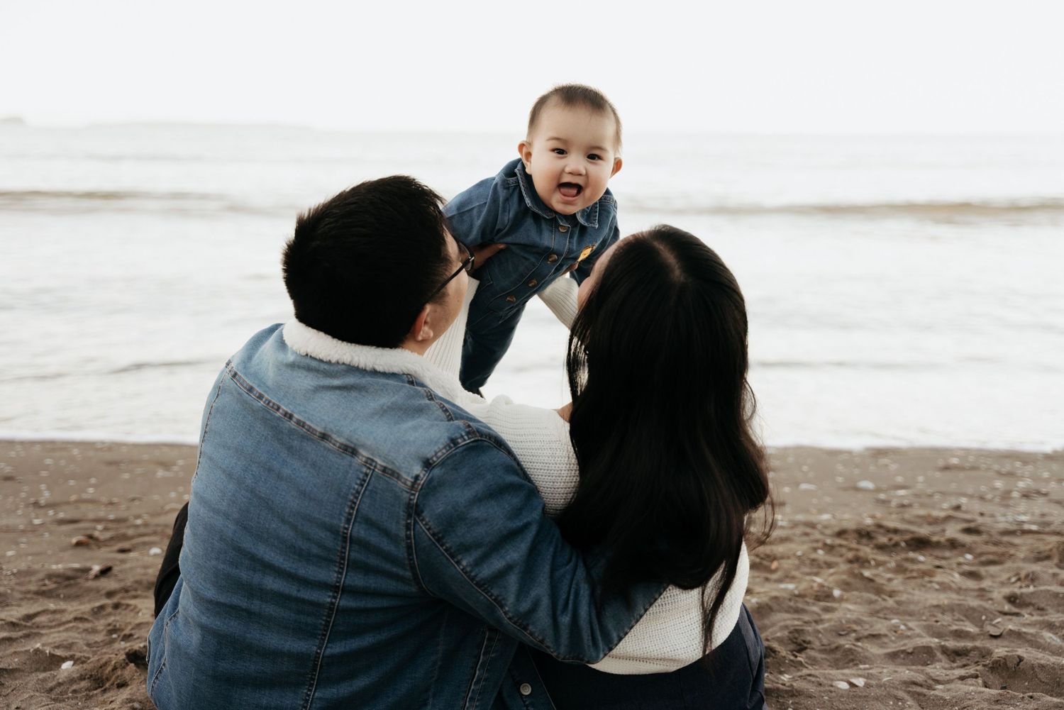 Parents sitting on a sandy beach holding up their laughing baby, with calm ocean waves in the background.