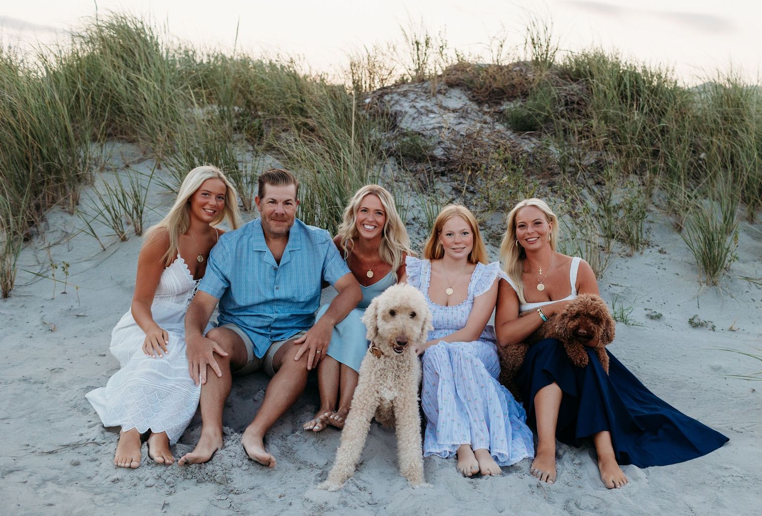 Family of five smiling on sandy beach dunes with a golden doodle and brown dog during golden hour.