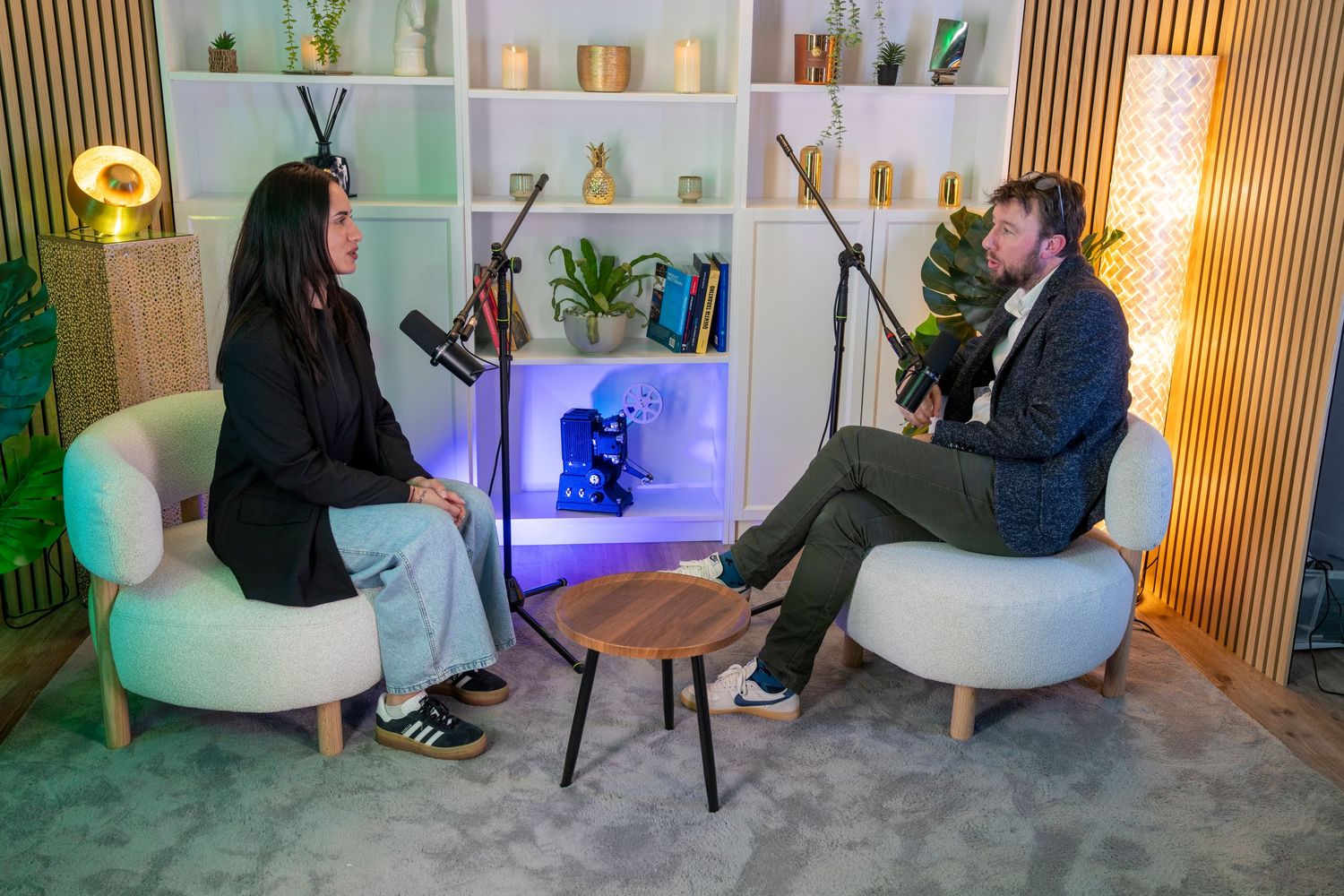 Two people sit in white chairs for a podcast recording session in a modern, plant-decorated studio with microphones.
