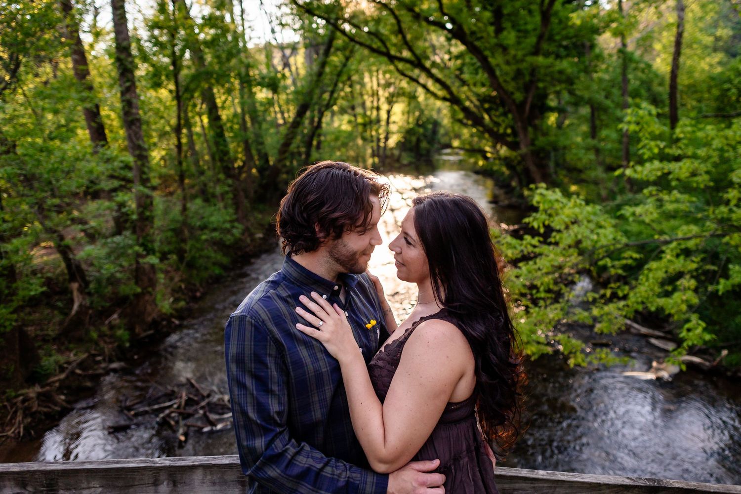 Couple embracing on a bridge surrounded by lush green trees and a flowing creek in a romantic outdoor setting.