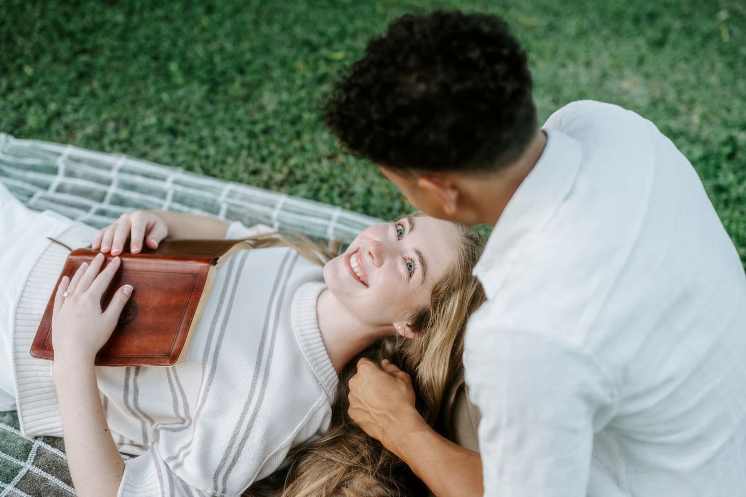 Young couple relaxing on a blanket outdoors, woman lying down smiling holding a book while man sits beside her.
