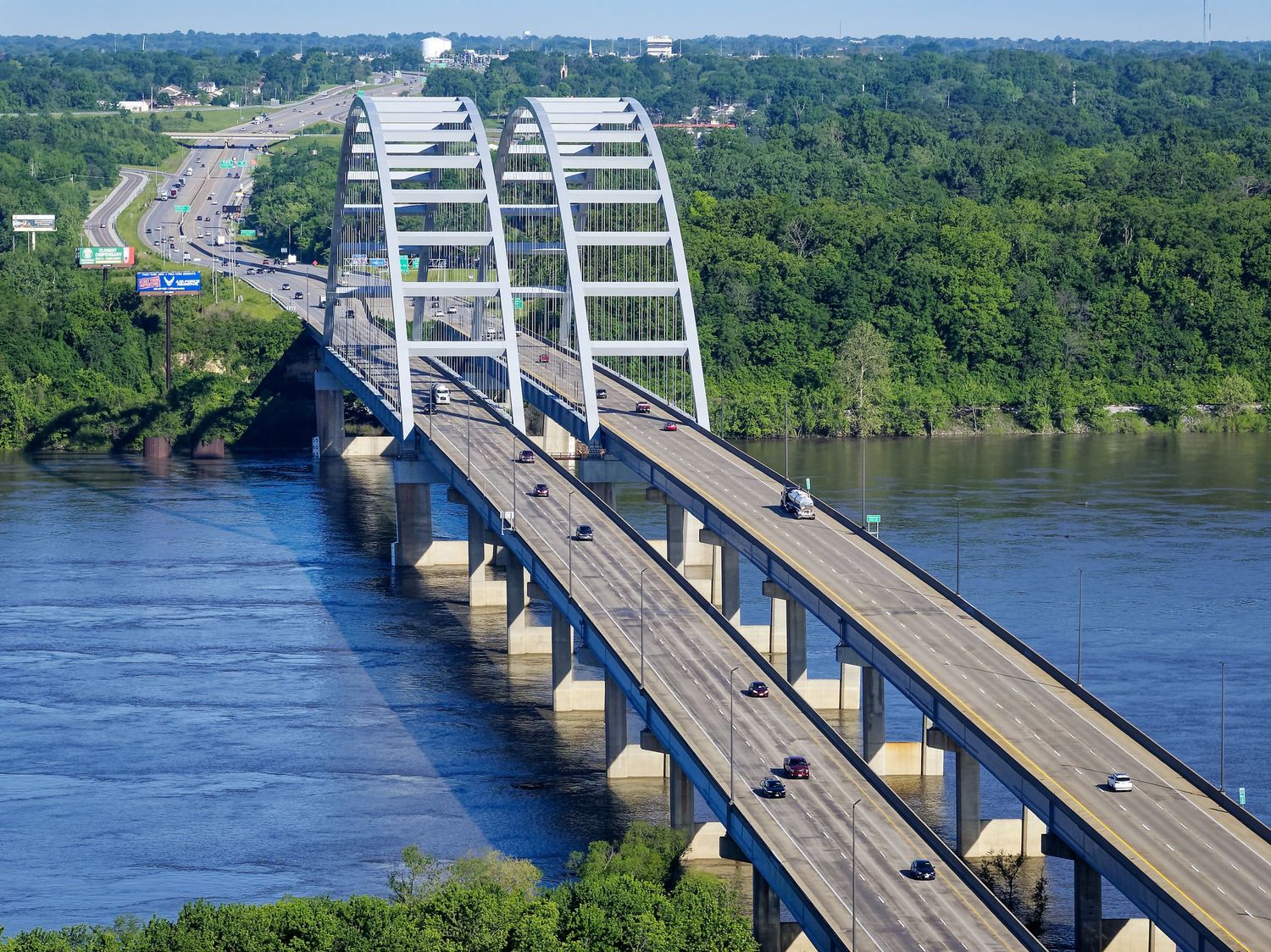 Aerial view of a modern cable-stayed bridge spanning a wide river with highway traffic surrounded by lush green trees.