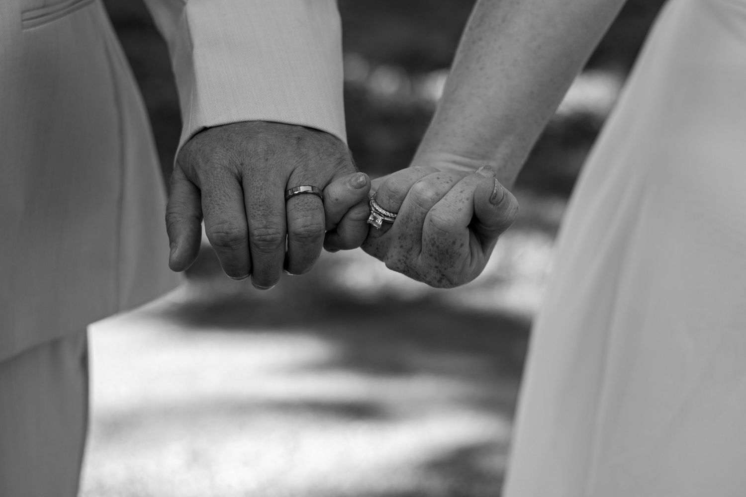 Black and white photo of a married couple holding hands, showing wedding rings, in elegant wedding attire.
