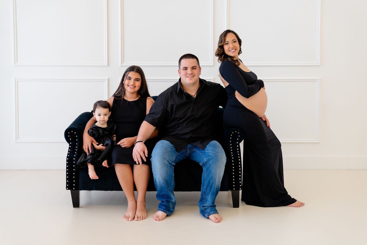 Family maternity photo with pregnant woman in black gown, man in black shirt, teen girl, and baby on navy sofa.