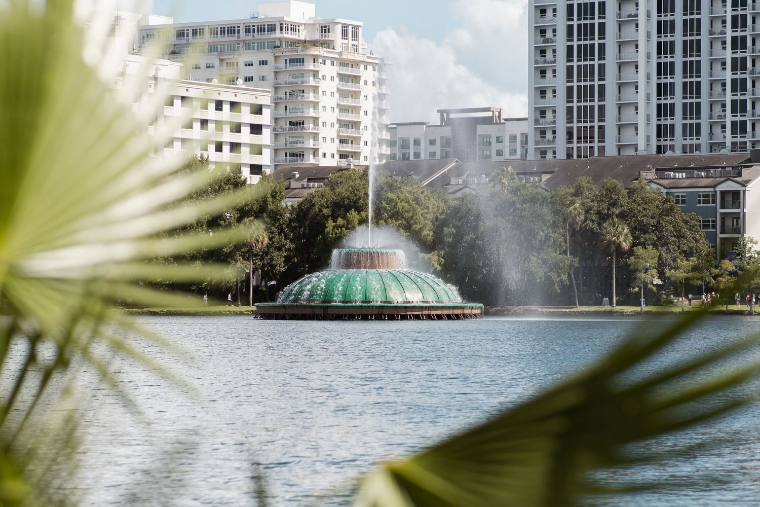 Green domed fountain spraying water on a lake surrounded by lush trees and modern city buildings in Orlando, Florida.
