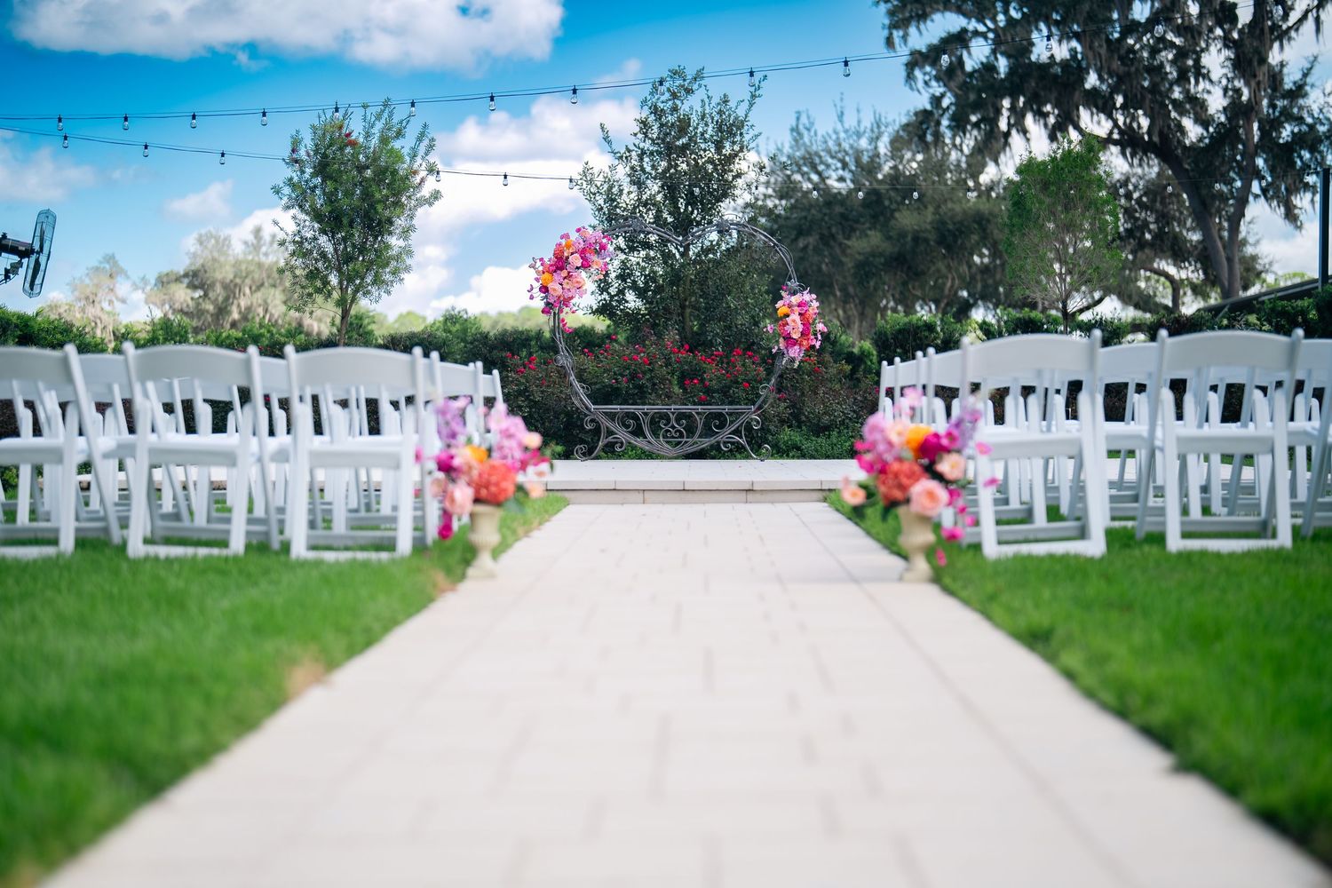 Outdoor wedding ceremony aisle with white chairs, pink floral arrangements, and a white runner on green grass under blue sky.