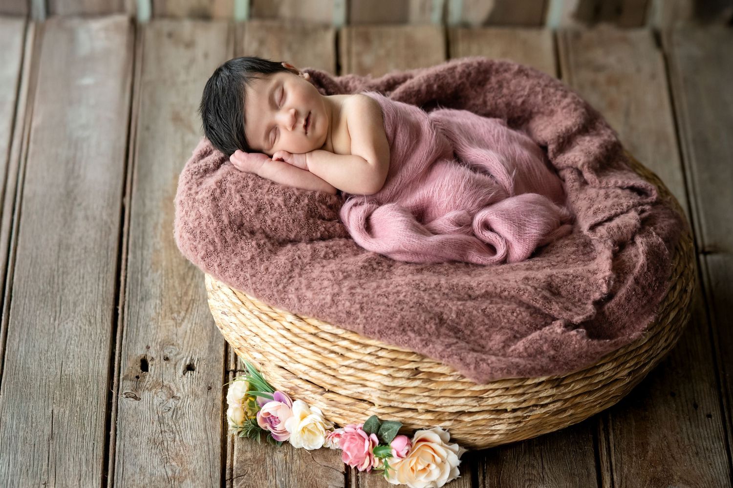Newborn baby sleeping peacefully in a wicker basket with pink fluffy blanket and floral decorations on wood floor.