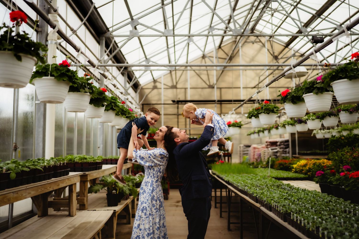 Family joyfully lifting children in a lush greenhouse filled with hanging flower baskets and green plants.
