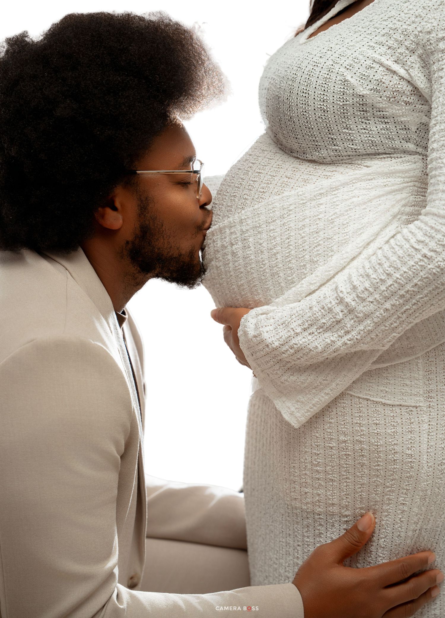 A man in a white suit kisses a pregnant woman's belly during a maternity photoshoot.
