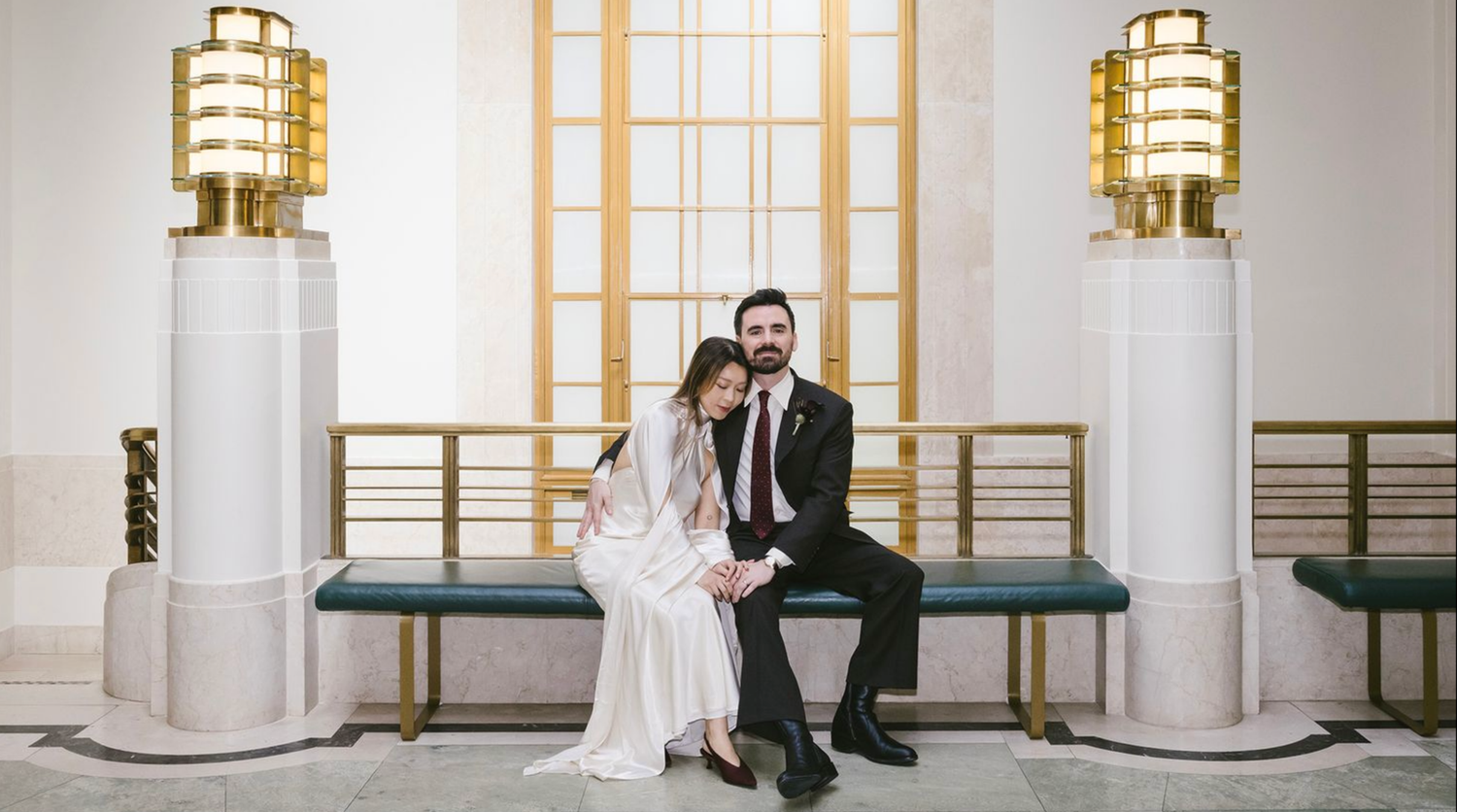 Couple walks hand-in-hand down aisle in elegant venue, bride in white jumpsuit, groom in plaid suit, guests applauding.