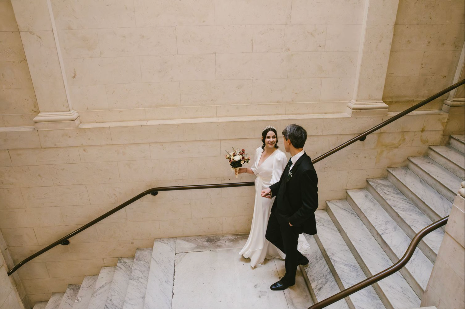 Bride and groom share a romantic moment on grand marble staircase, bride holding bouquet in elegant wedding attire.