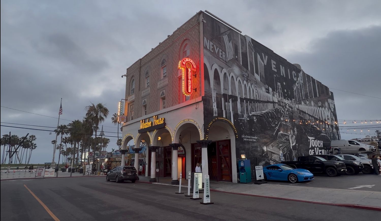 Historic hotel building with neon sign and large mural on exterior wall, parked cars on street under cloudy sky.