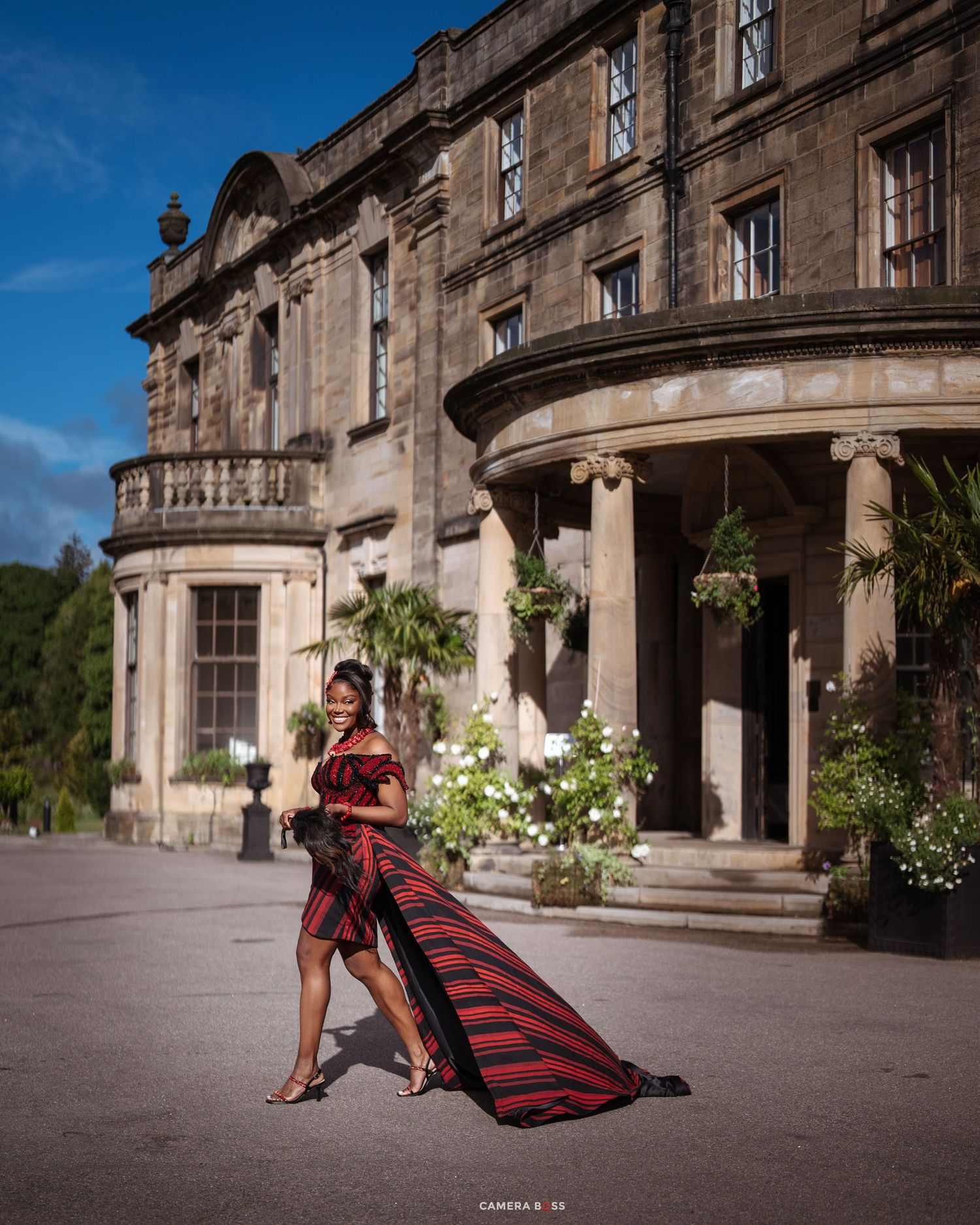 Woman in red and black striped gown posing before a grand historic stone mansion with columns and lush greenery.