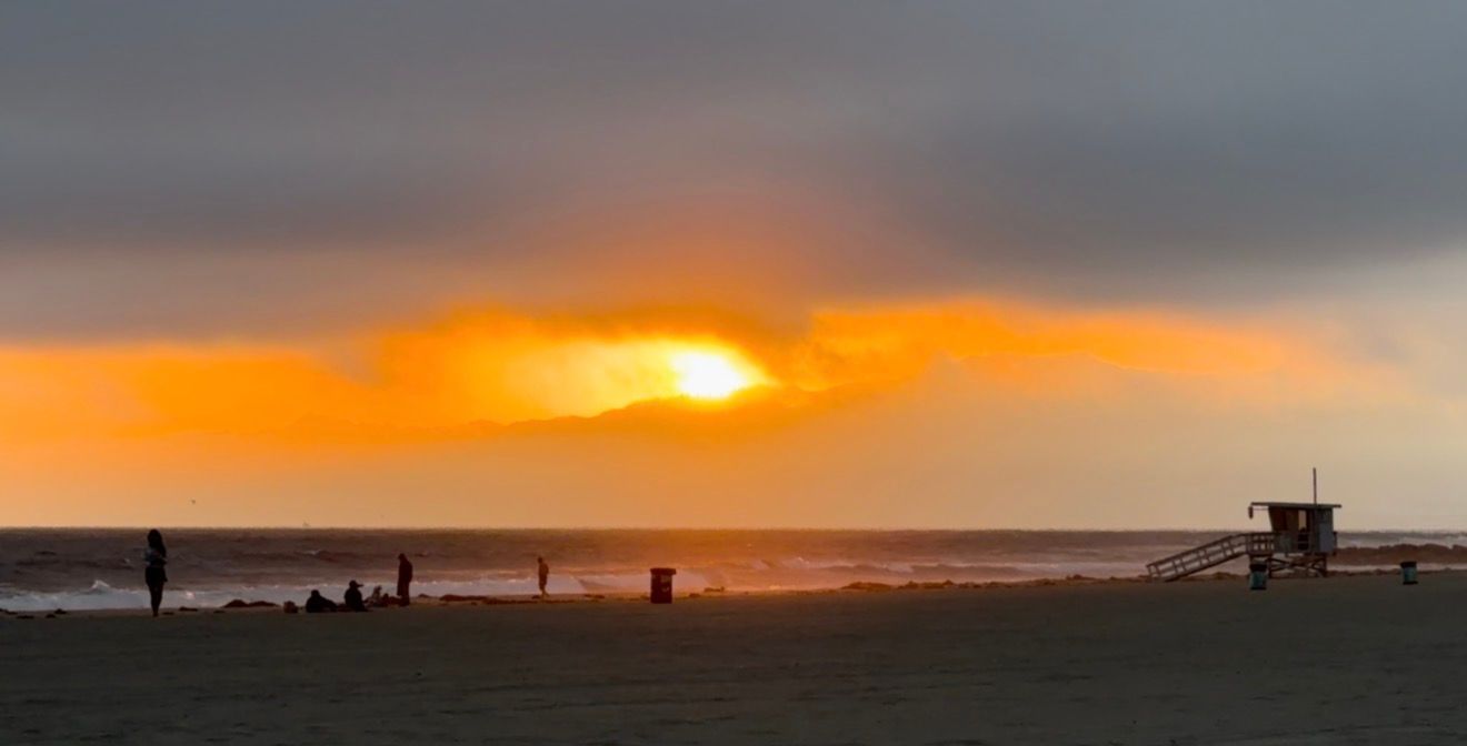 Dramatic sunset over a beach with silhouetted figures and a lifeguard tower under glowing orange skies.