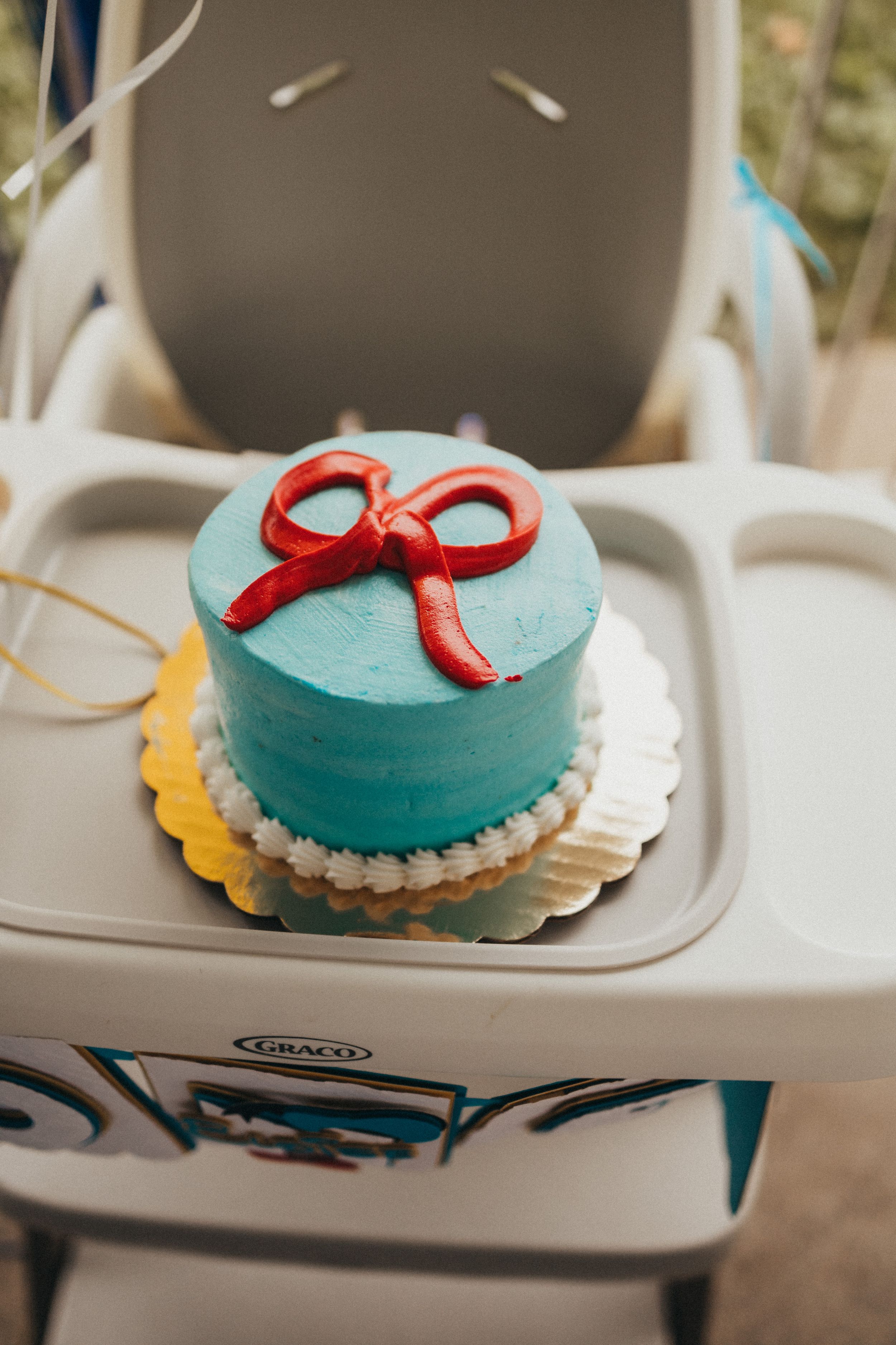 Teal smash cake with red fondant bow on white doily, displayed on Graco high chair tray for first birthday.