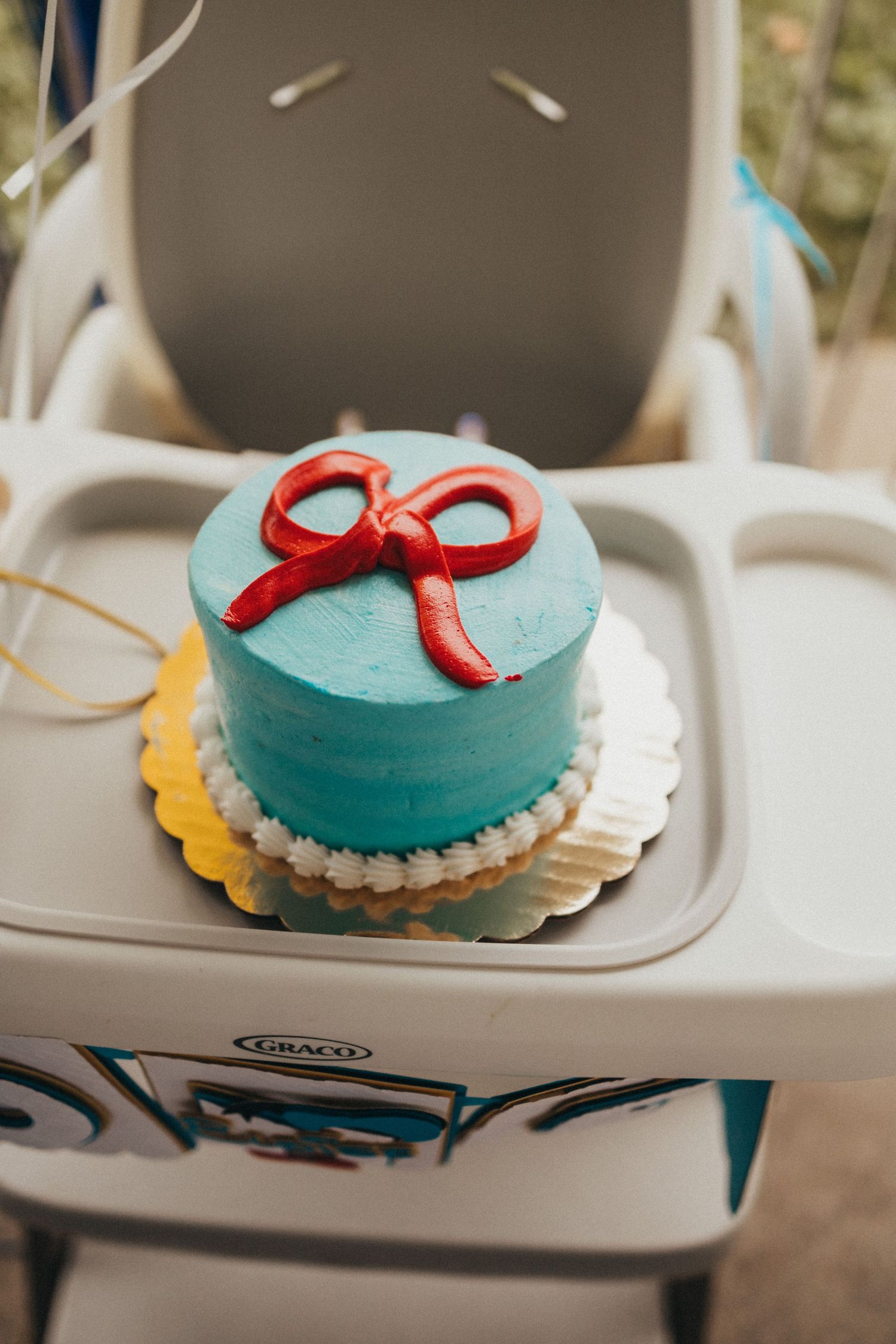 Teal smash cake with red fondant bow on white doily, displayed on Graco high chair tray for first birthday.