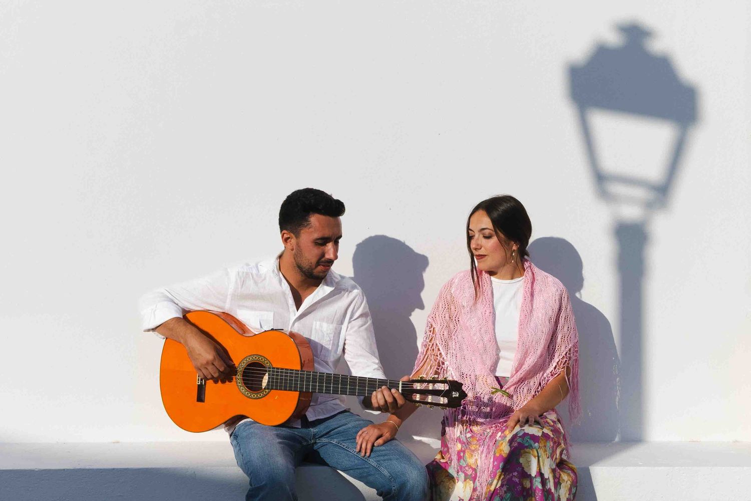 A woman in a pink shawl and a man playing an acoustic guitar stand against a white wall.
