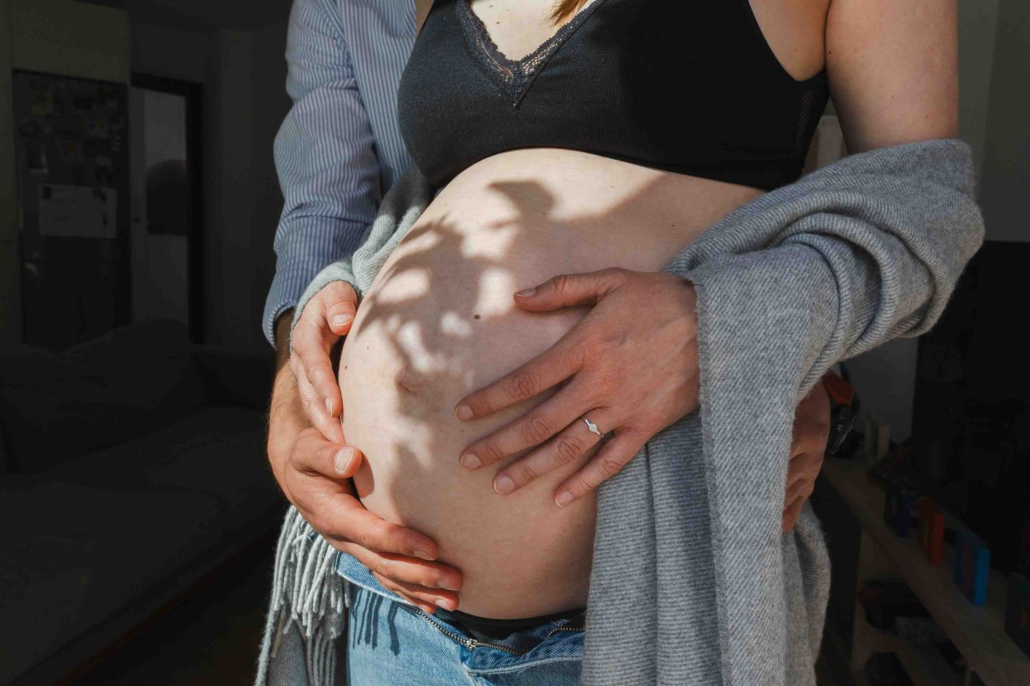 Pregnant woman in black bra and jeans with partner's hands cradling baby bump in warm sunlight.